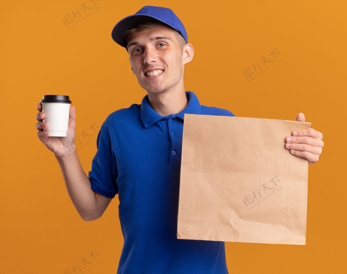 包装微笑着的年轻金发送货男孩拿着纸包和杯子橙色纸杯子