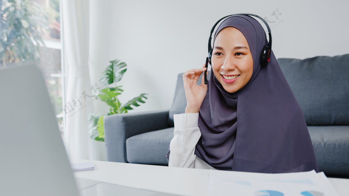 个人穆斯林女士戴着耳机使用笔记本电脑在客厅工作时 在会议视频通话中与同事谈论计划网络隔离自由职业者