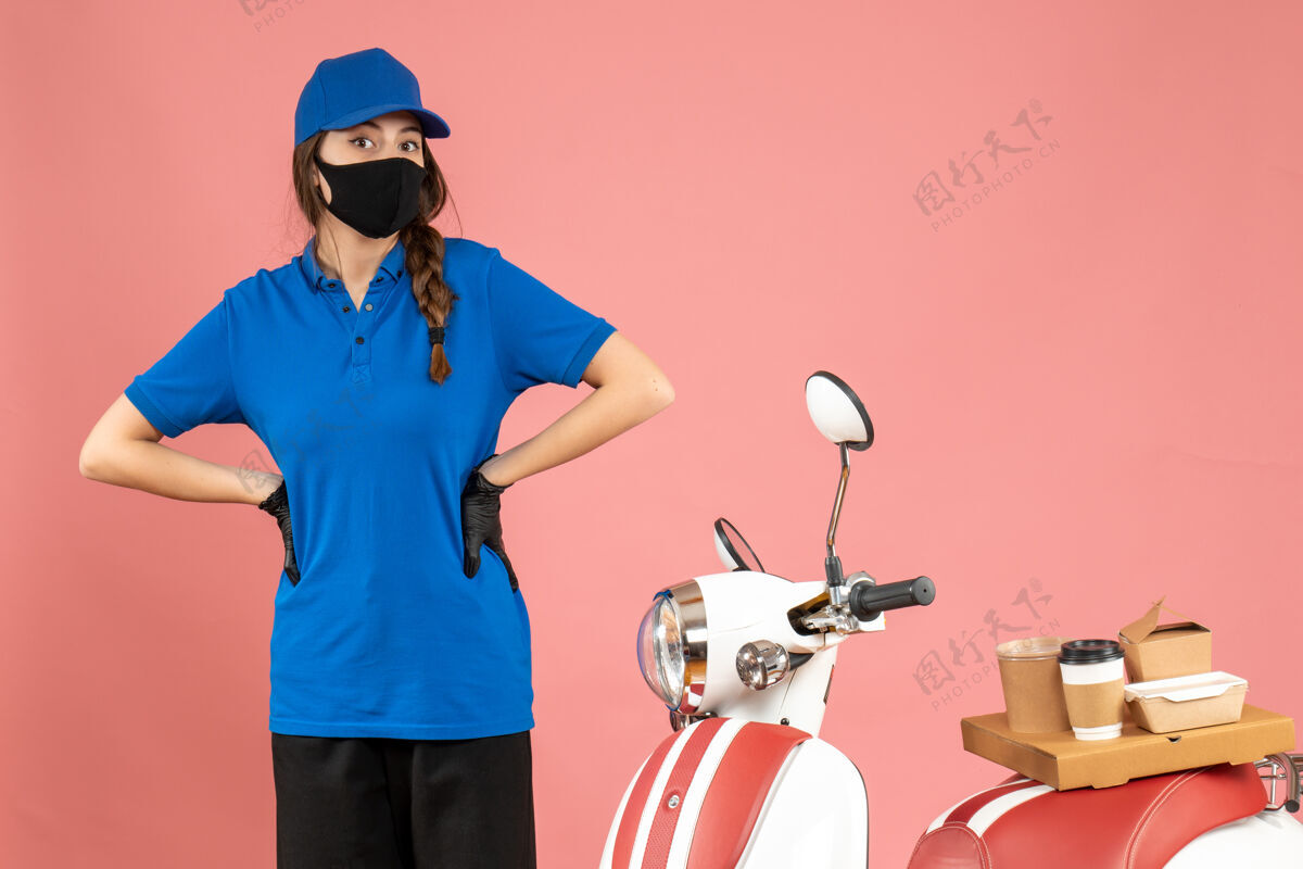 头盔顶视图混乱的快递女孩在医疗面具站在摩托车旁边与咖啡蛋糕上的粉彩桃色背景蛋糕困惑工人