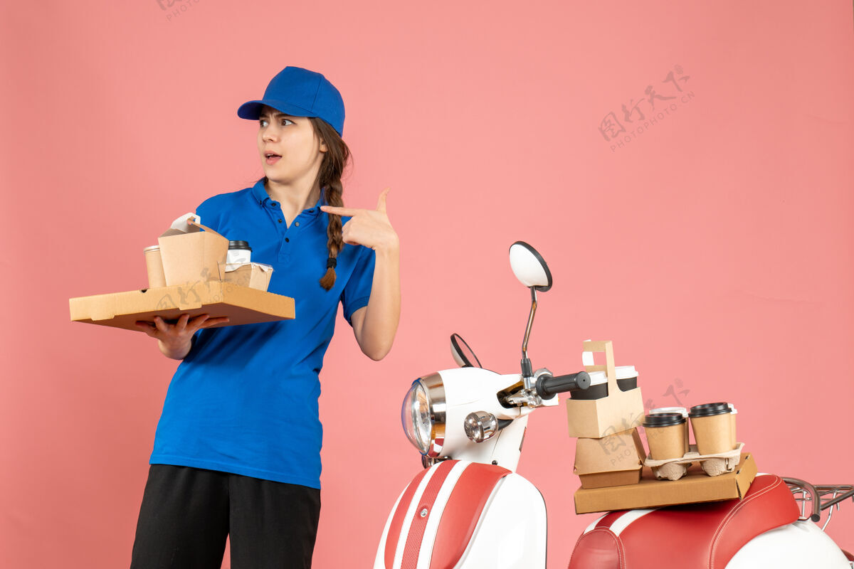 背景前视图好奇的信使女士站在摩托车旁边拿着咖啡和小蛋糕粉彩桃色背景工作男桃