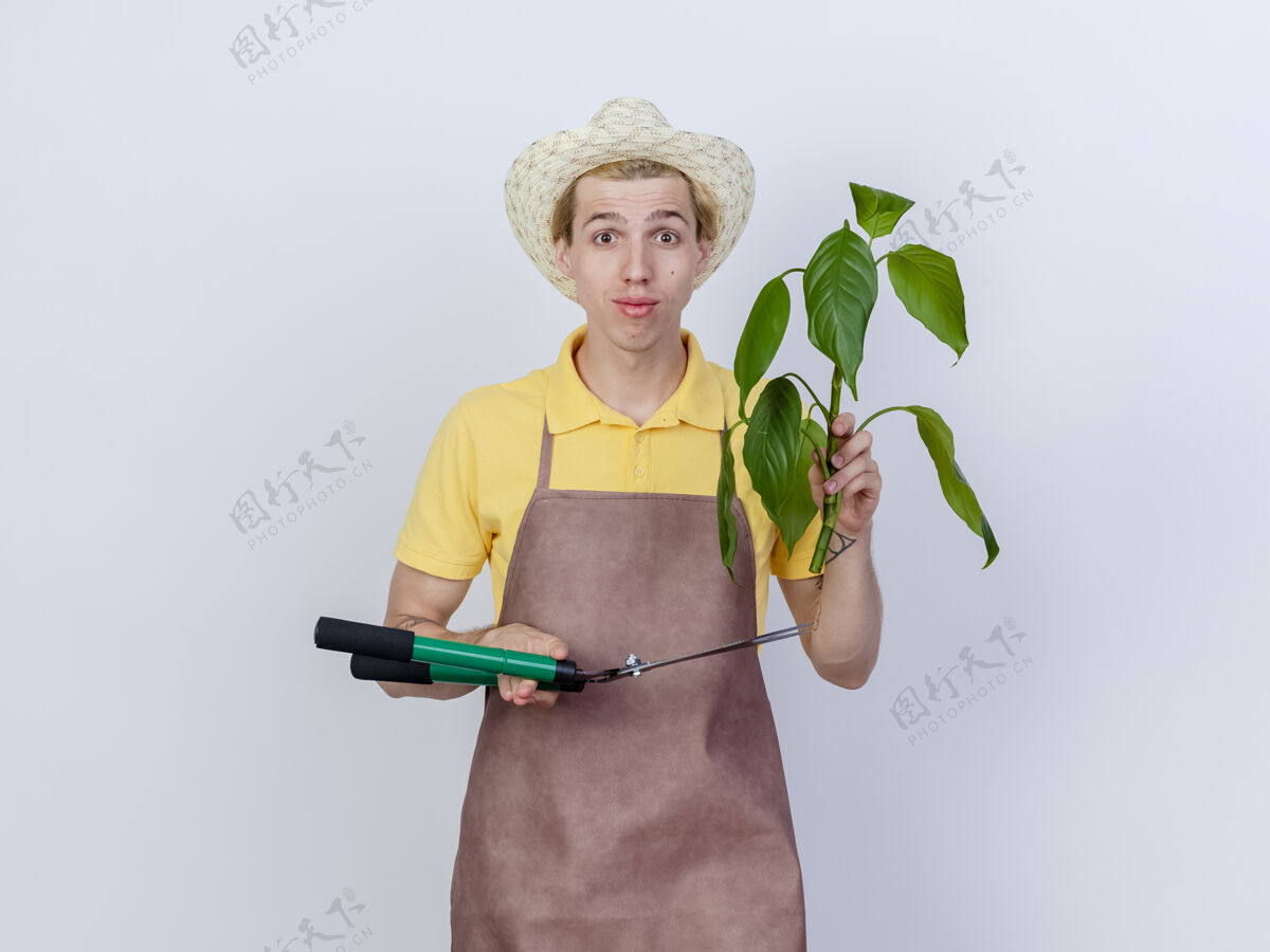 花园年轻的园丁 穿着连体衣 戴着帽子 拿着植物和篱笆剪 脸上带着微笑男人站着剪子