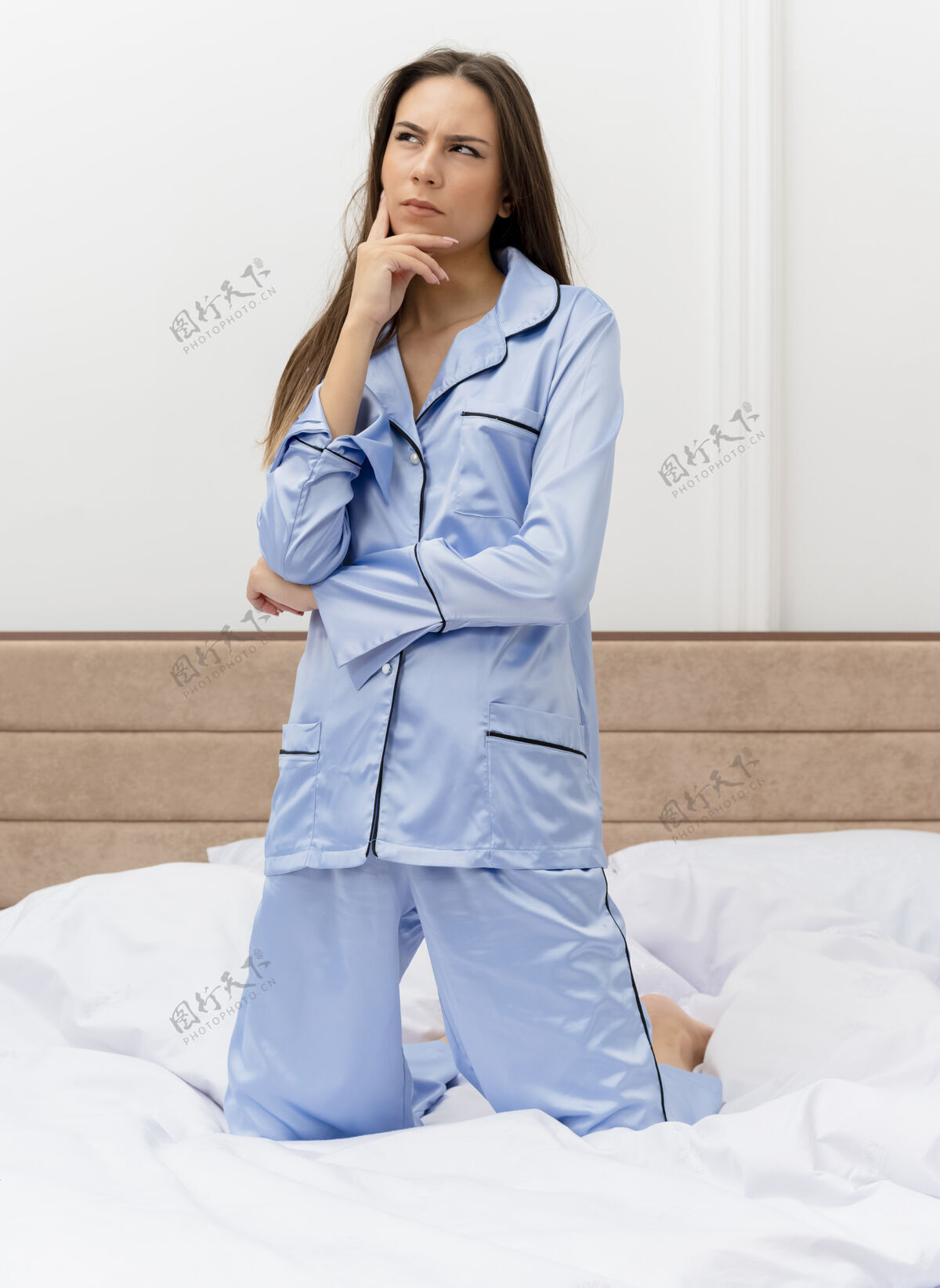 坐着穿着蓝色睡衣的年轻漂亮女人坐在床上 困惑地看着卧室的内部床室内漂亮