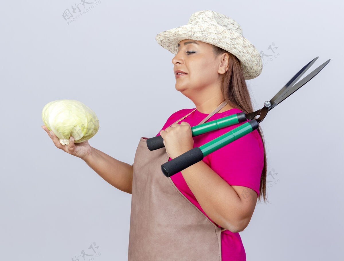 花园围裙戴帽子的中年园丁妇女拿着卷心菜和树篱剪看着卷心菜 脸上带着微笑脸抱站