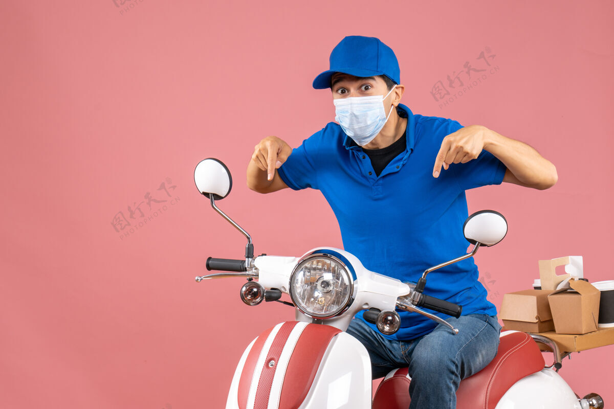 车辆前视图的快递员戴着医用面罩 戴着帽子坐在滑板车上 指着粉色背景背景面具指向