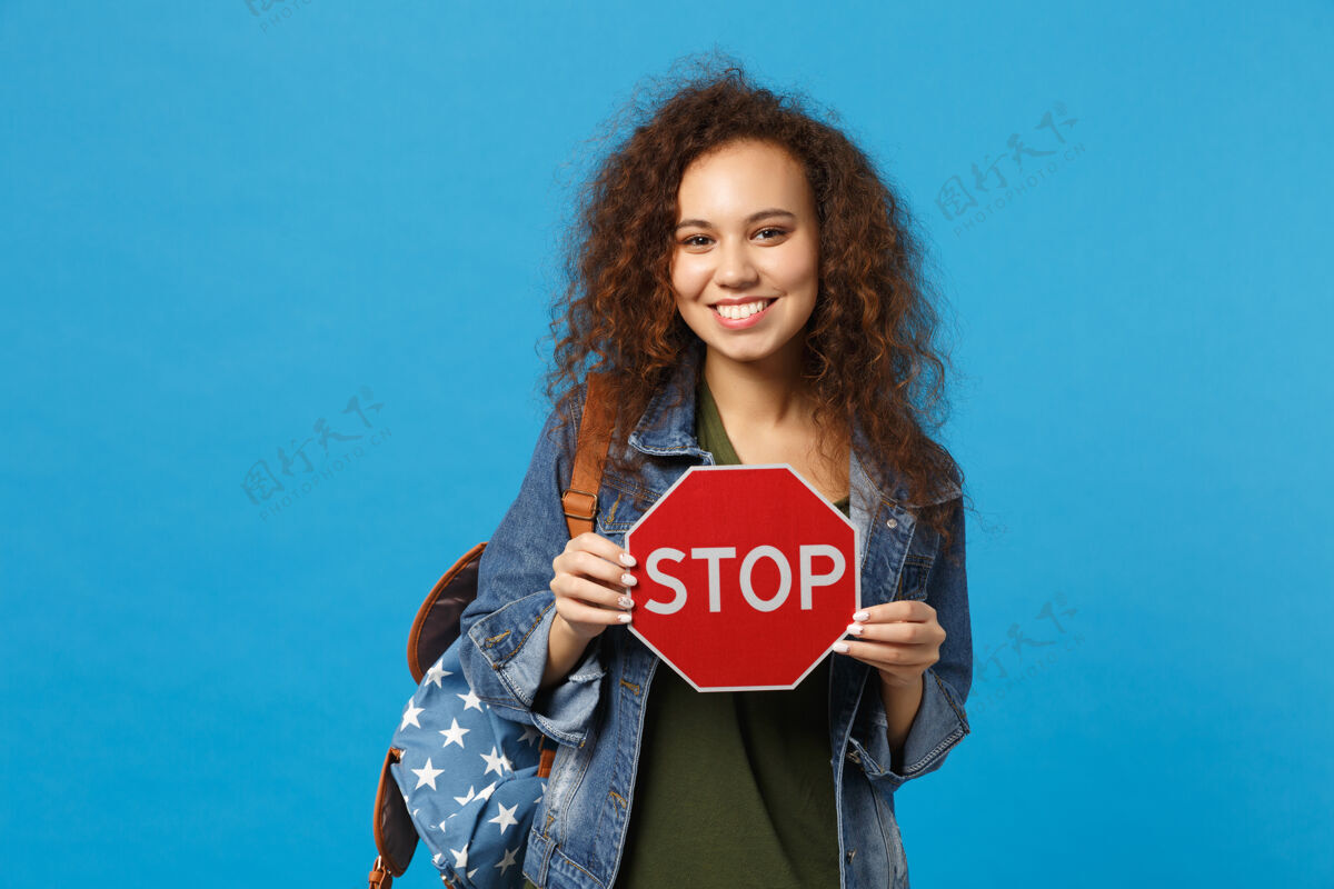 站立年轻的非洲裔美国女孩十几岁的学生在牛仔服装 背包举行停止孤立的蓝色墙壁显示停止红色