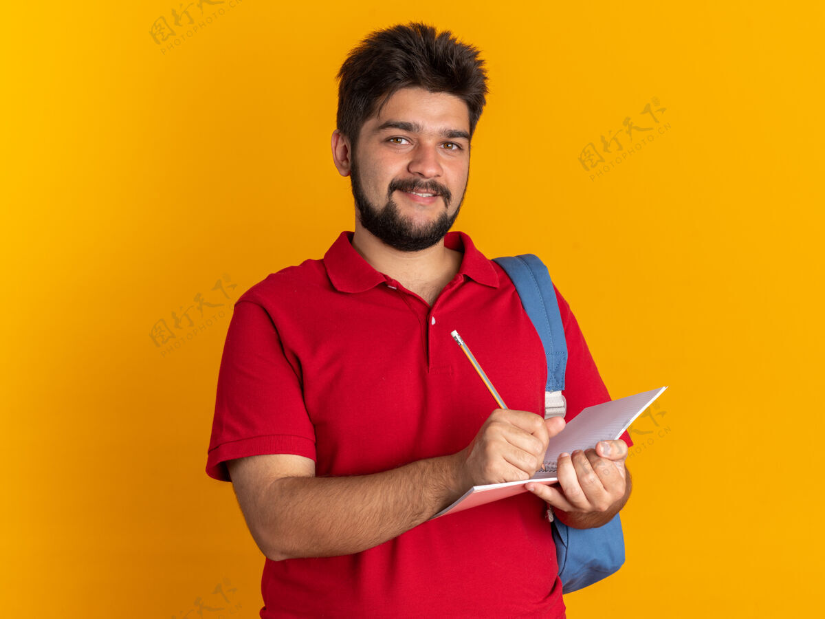马球快乐的年轻留着胡子的学生 穿着红色马球衫 背着背包 拿着笔记本和铅笔 站在橘色的墙上 开心地微笑着笔记本姿势写作