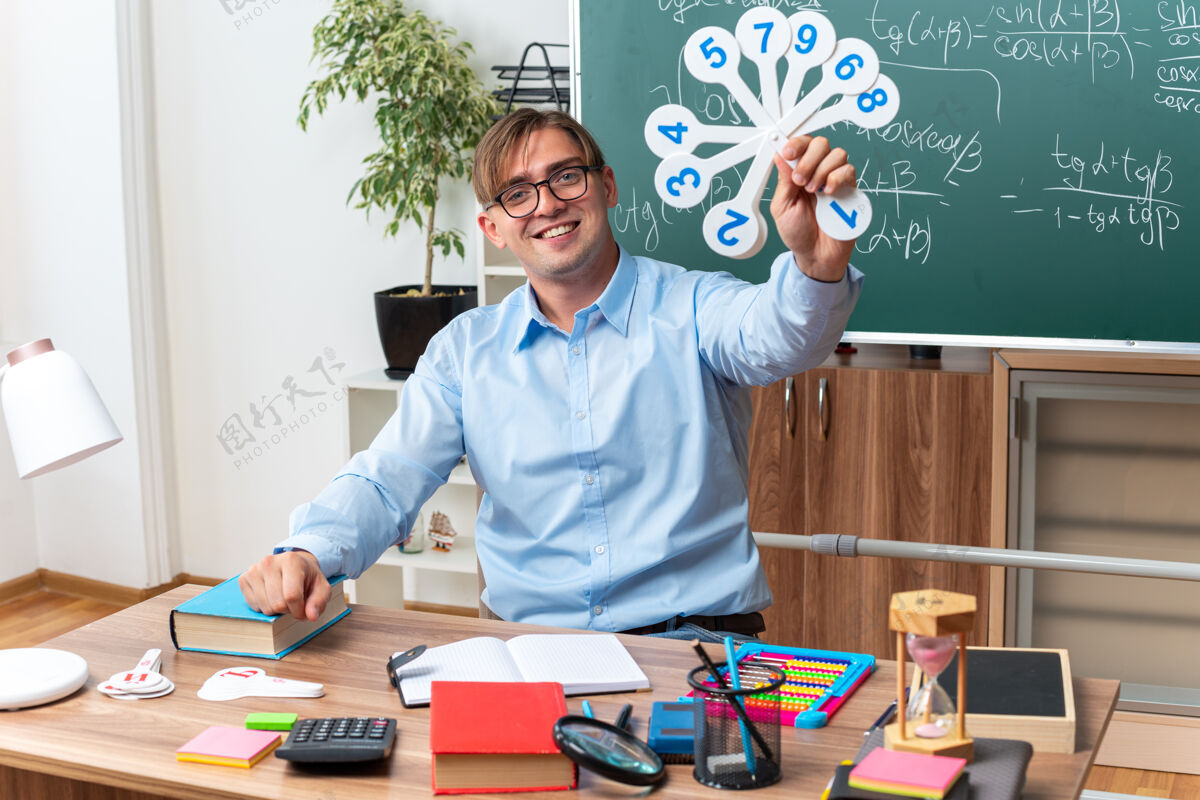 课桌年轻的男老师戴着眼镜 展示着解释课的车牌 微笑着坐在课桌旁 教室里的黑板前放着书和笔记课盘子展示