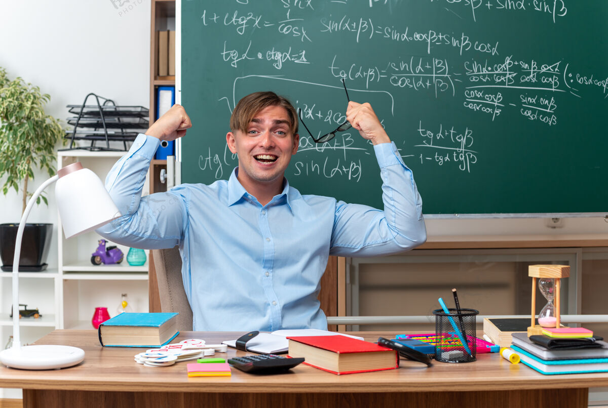 兴奋年轻的男老师举着眼镜 兴高采烈地举起拳头坐在课桌旁 手里拿着书和笔记 在教室的黑板前黑板教室课桌