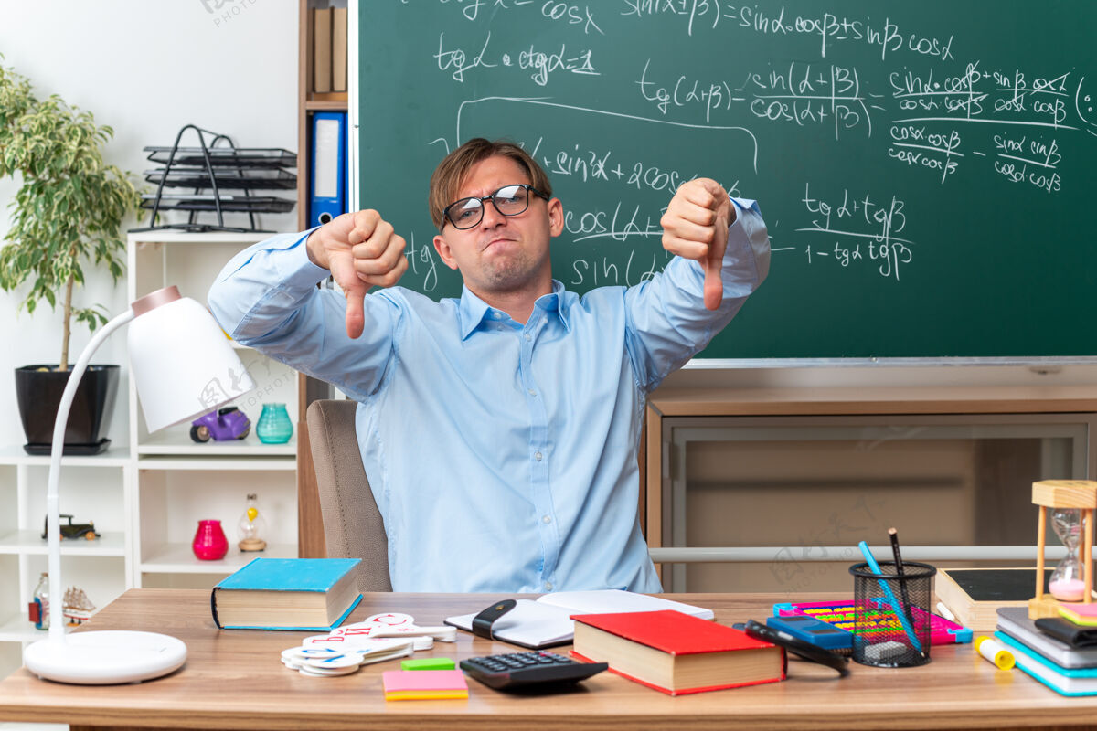 穿戴眼镜的年轻男老师不高兴地竖起大拇指坐在课桌旁 教室的黑板前放着书和笔记笔记不高兴拇指