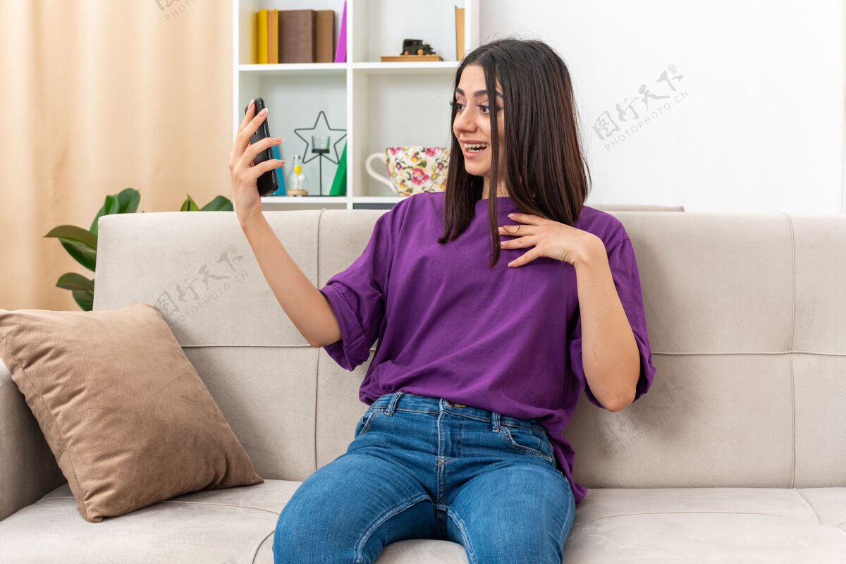 积极穿着休闲服的年轻女孩拿着智能手机打视频电话快乐而积极的微笑自信地坐在客厅的沙发上视频光生活