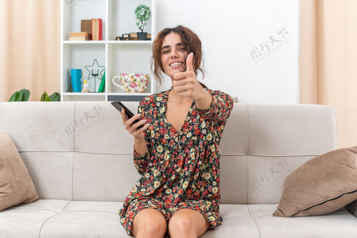 生活在明亮的客厅里 一个穿着花裙子的年轻女孩坐在沙发上 手里拿着智能手机 兴高采烈地笑着竖起大拇指欢呼花卉服装