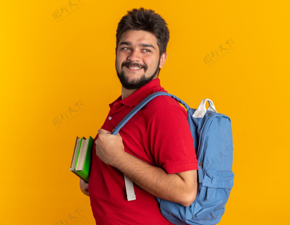 胡子年轻的留着胡子的学生 穿着红色马球衫 背着背包 拿着笔记本 面带微笑 自信 快乐 积极地站在橙色的墙上年轻站靠边
