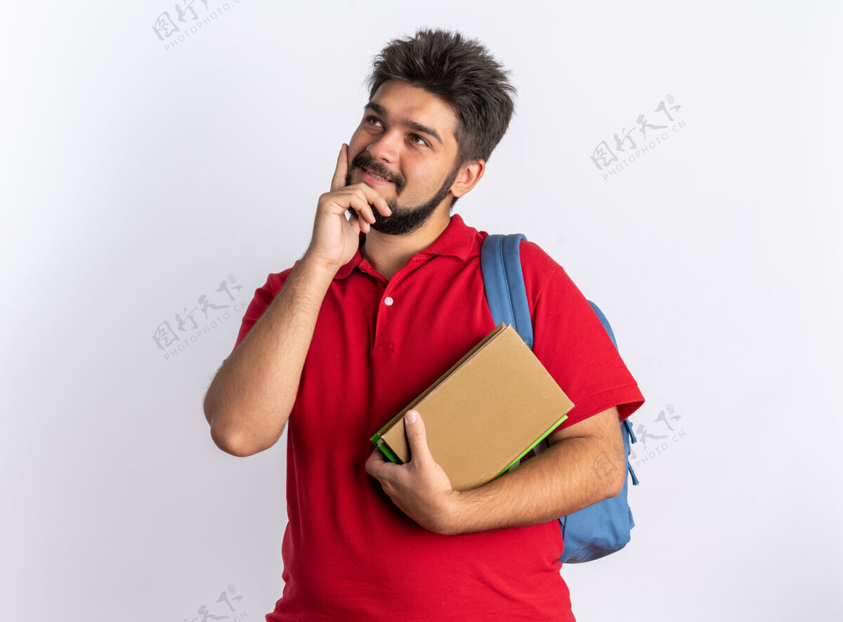 思考年轻的留着胡子的学生 穿着红色马球衫 背着背包 拿着笔记本 面带微笑 抬头看着 站在白墙上思考着积极的事情手势背包马球