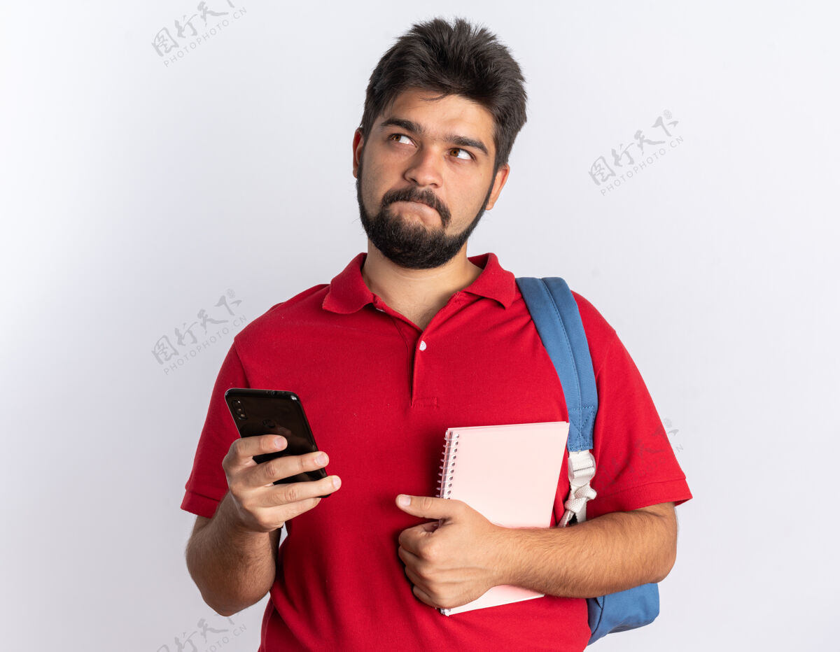 马球一个留着胡子的年轻学生 穿着红色马球衫 背着背包 手里拿着智能手机和笔记本 站在白色的墙上困惑地看着我人胡须笔记本