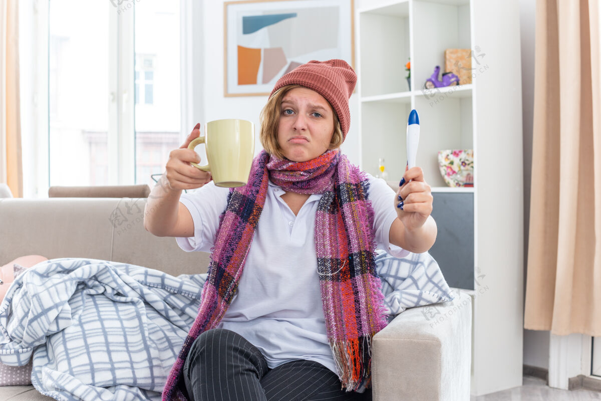周围不健康的年轻女子戴着暖和的帽子 脖子上围着围巾 手里拿着一杯茶和温度计 坐在明亮的客厅里的沙发上 看起来很担心温暖担心温度计