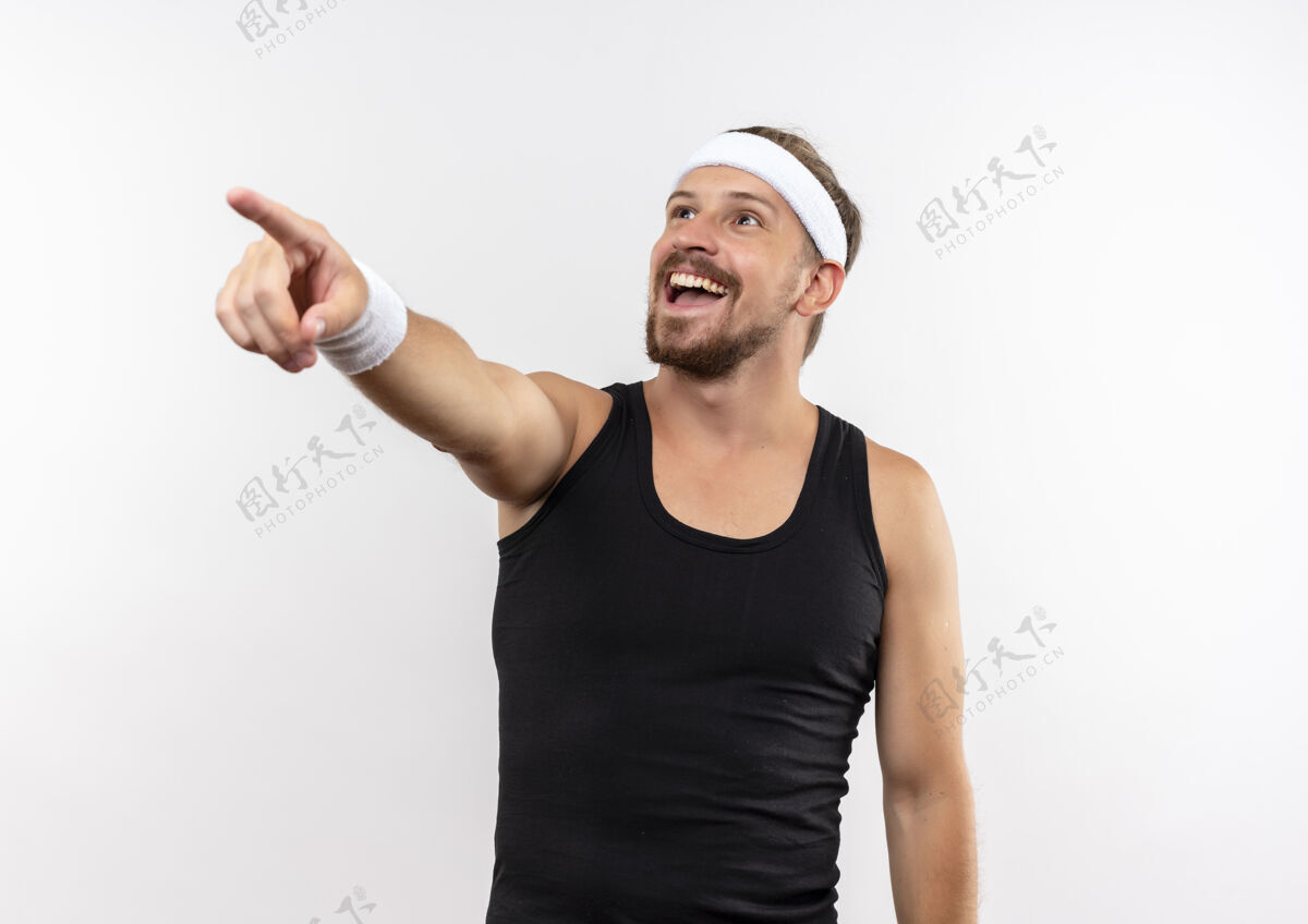 欢乐快乐的年轻帅气的运动型男人戴着头带和腕带 指着和看着隔离在白色墙壁上的一面 复制空间男人英俊头带