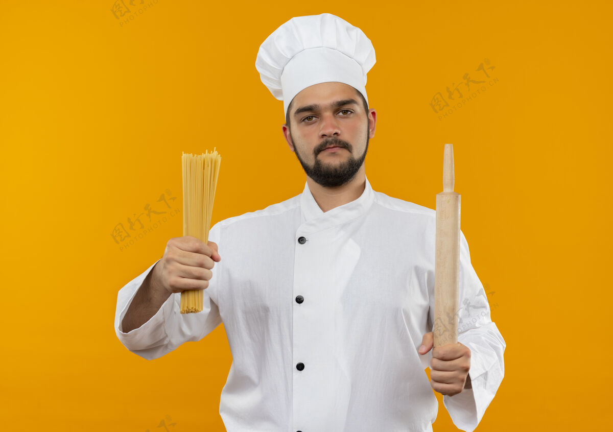 持有自信的年轻男性厨师身着厨师制服 手拿意大利面和擀面杖 隔离在橙色的墙上信心卷别针