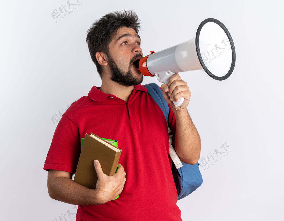 背包情绪激动的留着胡子的年轻学生 穿着红色马球衫 背着背包 拿着笔记本 对着站在白墙上的扩音器大叫人胡子年轻人