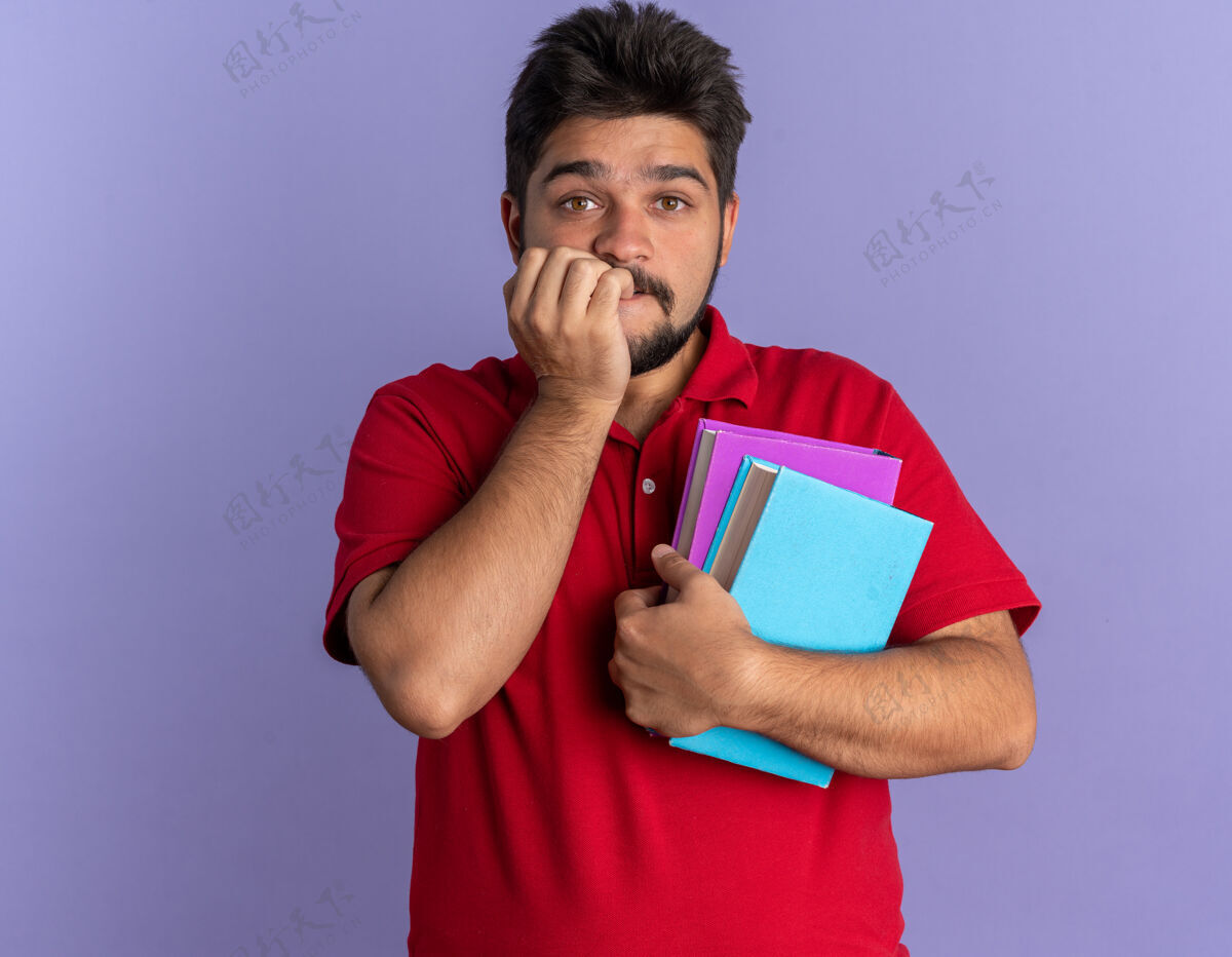 人年轻的留着胡子的学生 穿着红色马球衫 手里拿着书 紧张地咬着钉子站在蓝色的墙上马球姿势手势