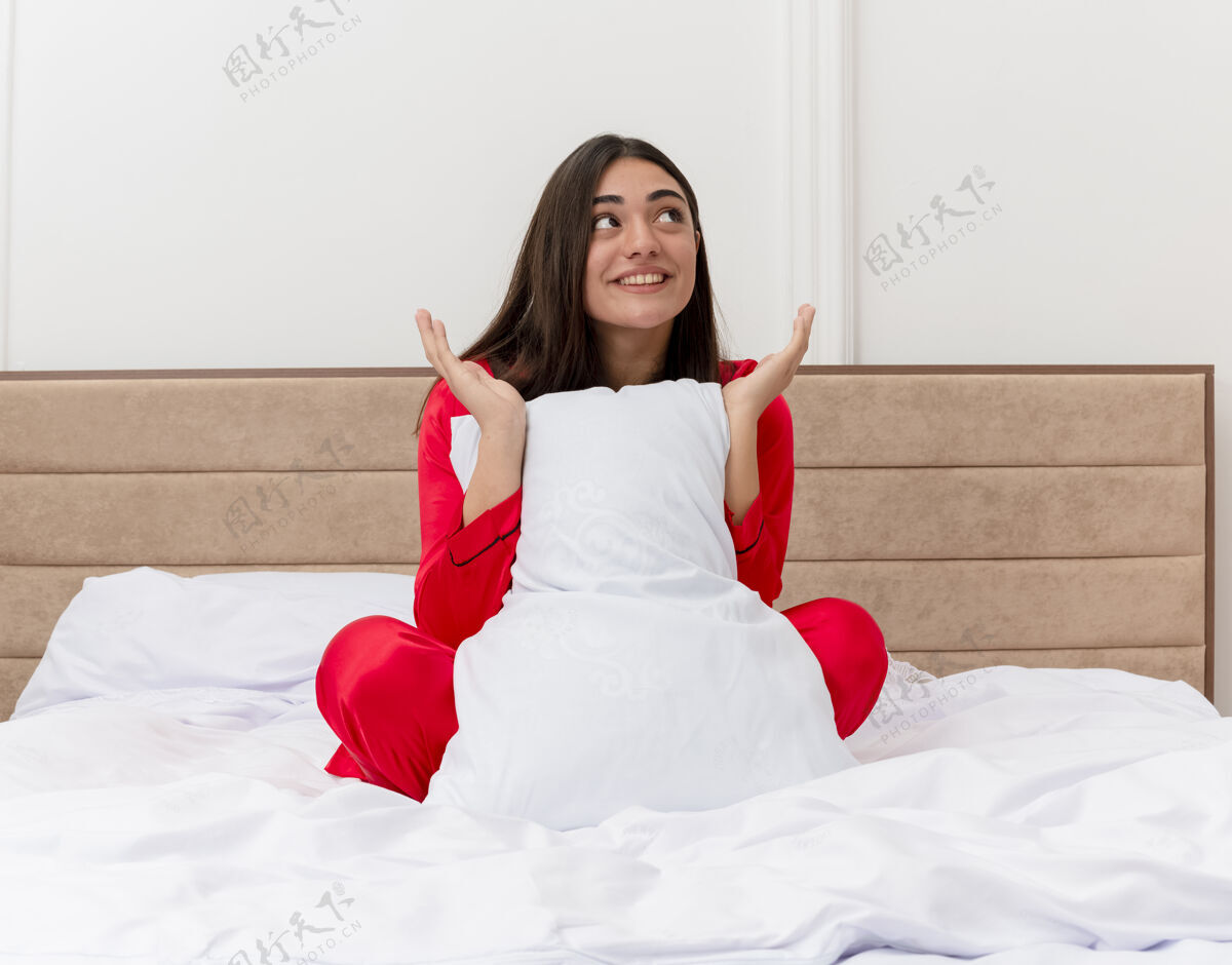 光穿着红色睡衣的年轻漂亮女人坐在床上 枕着枕头 面带微笑 在卧室里享受周末红枕头室内