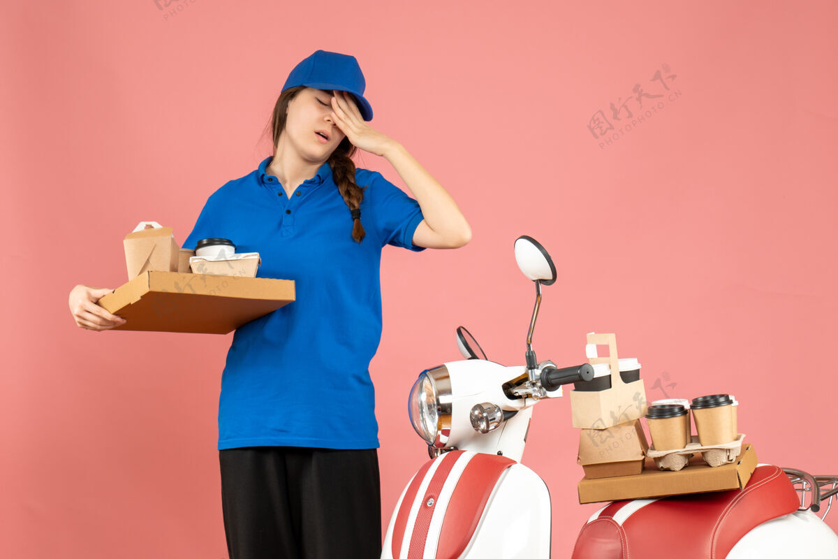 桃子疲惫的快递员女孩站在摩托车旁 手里拿着咖啡和小蛋糕 背景是柔和的桃色专业小成人
