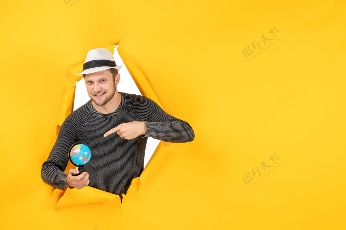 男孩一个微笑的男人 戴着帽子 手里拿着一个小地球仪 指着一面撕破的黄色墙上的一个小地球仪帽子地球仪孩子