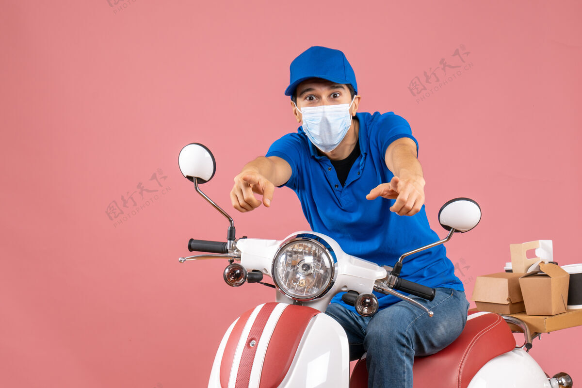 粉彩前视图自信的快递员戴着医用面罩戴着帽子坐在粉彩桃色背景的踏板车上快递员男车辆