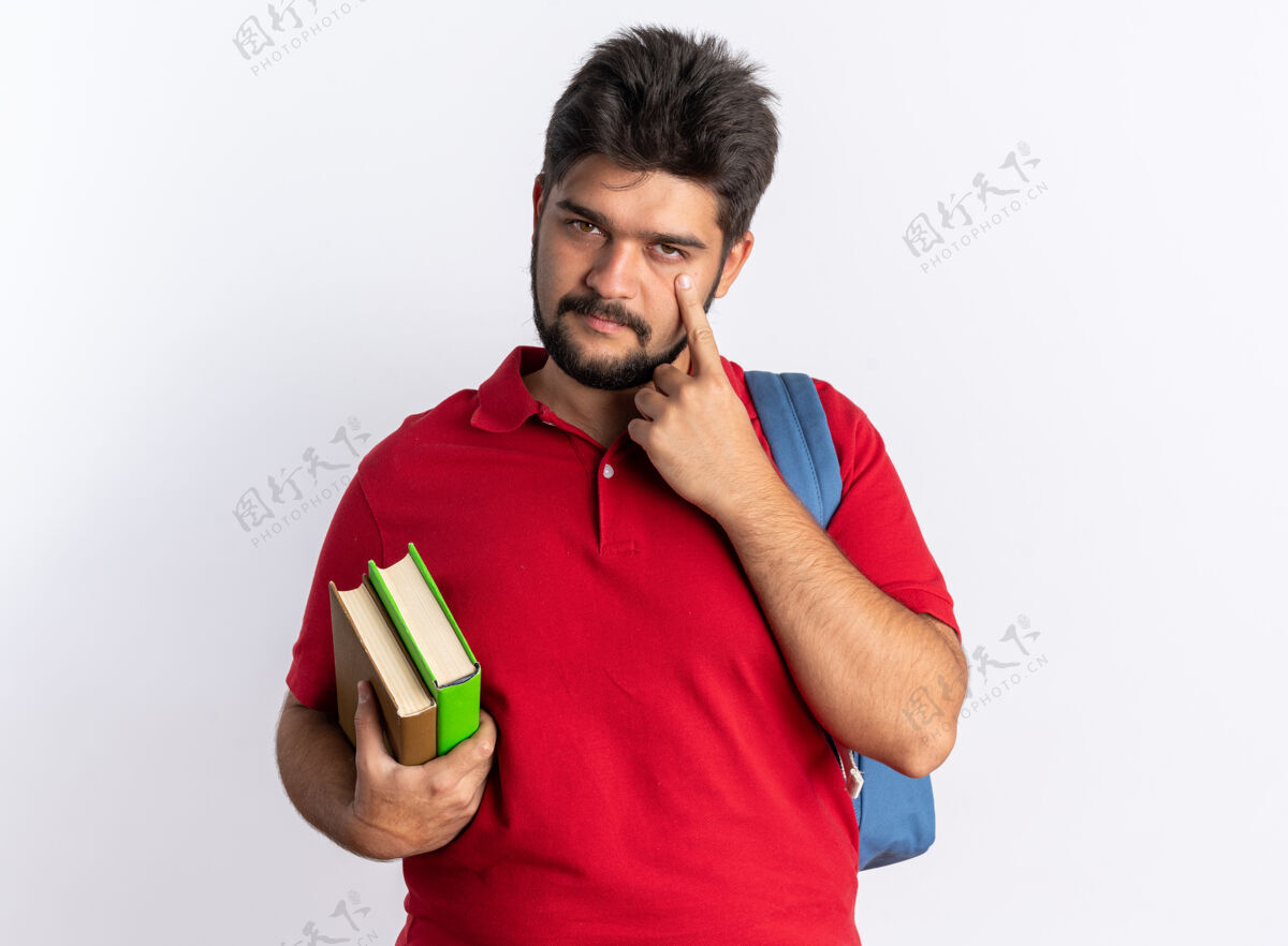 胡子年轻的留着胡子的学生 穿着红色马球衫 背着书包 拿着笔记本 用手指着他站在白墙上的眼睛马球年轻人姿势