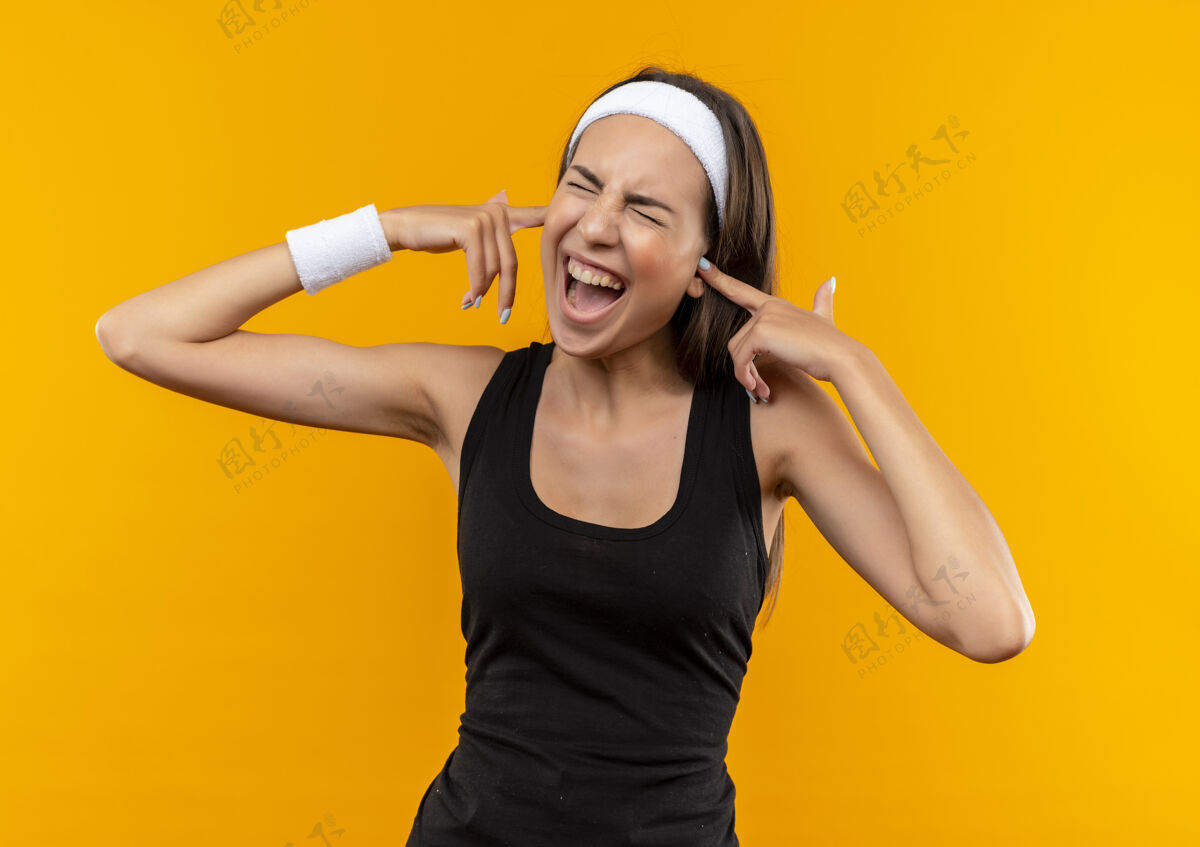 女孩愤怒的年轻漂亮的运动女孩戴着头带和腕带把手指放在耳朵里闭着眼睛孤立在橙色的墙上运动年轻腕带
