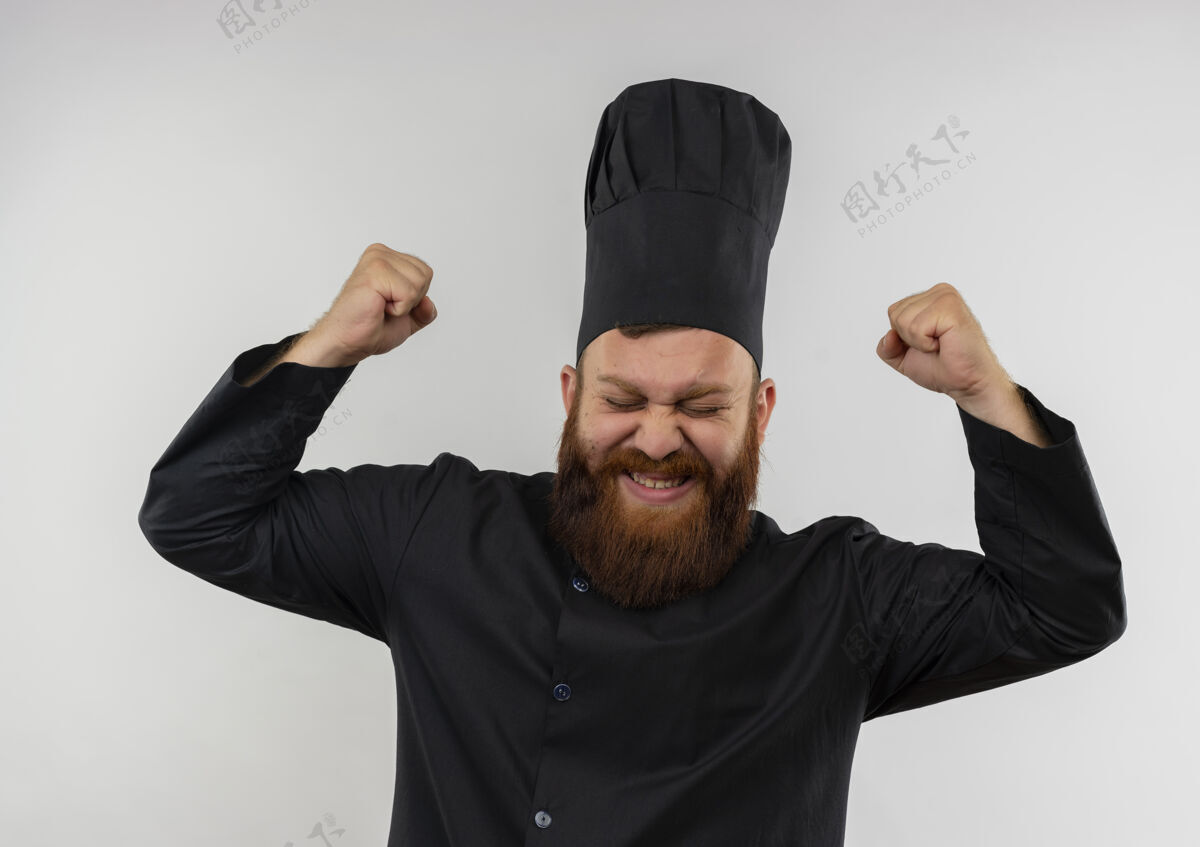 提高快乐的年轻帅哥厨师身着厨师制服举起拳头闭上眼睛孤立在白墙上厨师帅气烹饪