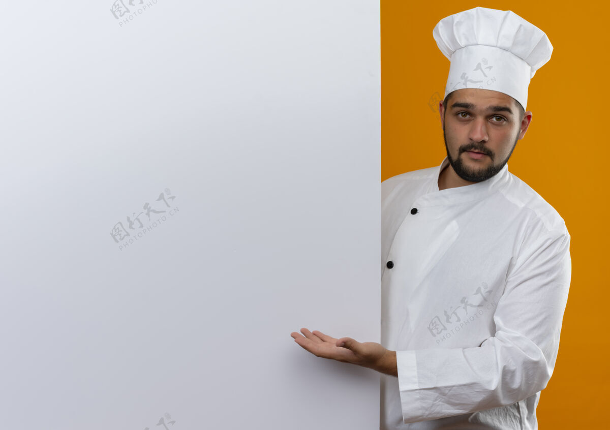 制服穿着厨师制服的年轻男厨师站在身后 用手指着白色的墙壁 看起来孤立在橙色的墙上 留有复印空间指向手烹饪