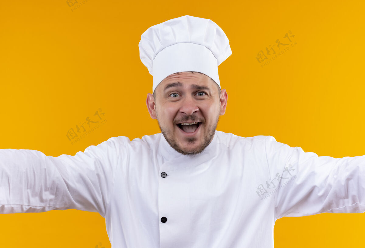烹饪年轻帅气的厨师身着厨师制服 张开双臂 孤零零地站在橙色的墙上开放厨师制服