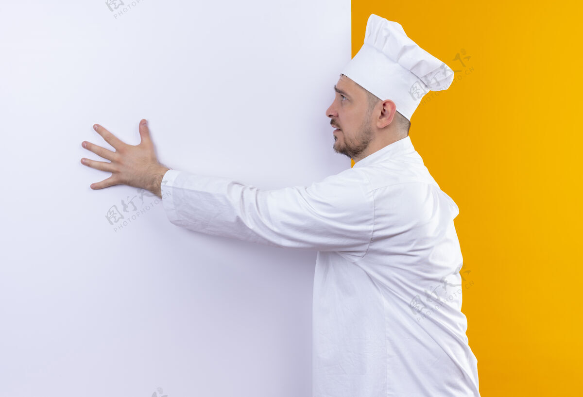 墙年轻帅气的厨师穿着厨师制服站在白色的墙前 把手放在上面 看着橙色墙上孤立的一面印象壁板厨师