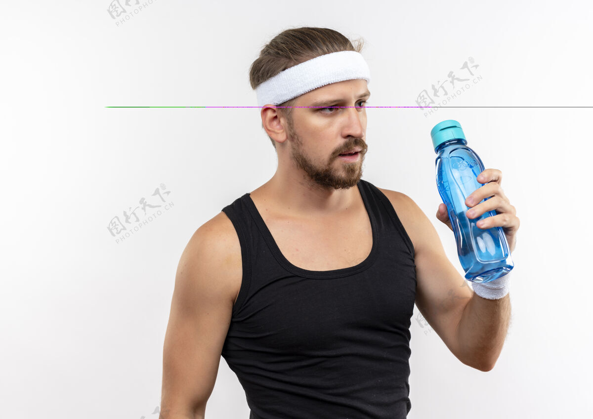 运动自信的年轻帅气的运动型男人戴着头带和腕带拿着水瓶隔离在白色的墙上帅气戴年轻