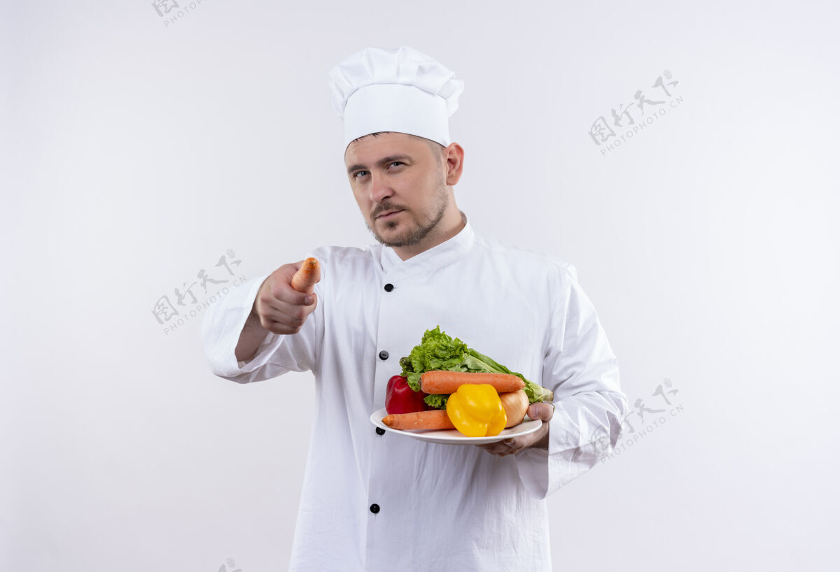 胡萝卜自信的年轻帅哥厨师穿着厨师制服拿着盘子里的蔬菜和胡萝卜指着孤立的白色墙壁拿着指着帅气