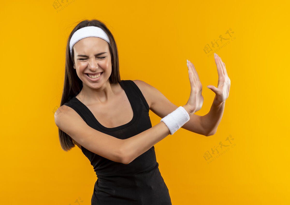 运动恼怒的年轻漂亮的运动女孩戴着头带和腕带 闭着眼睛 伸出双手孤立在橙色的墙上 不在旁边做手势伸展漂亮壁板