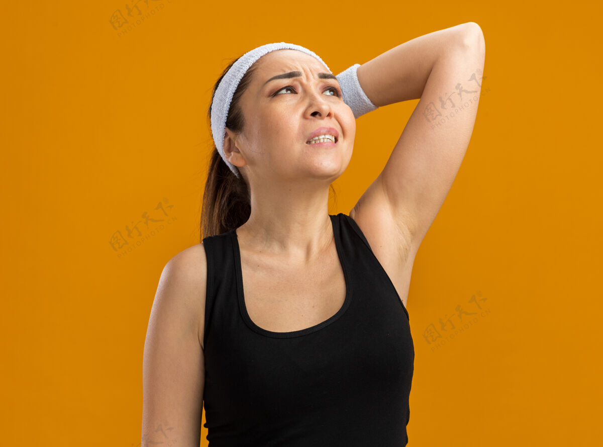 头带戴着头巾和臂章的年轻健身女士站在橘色的墙上 困惑地看着疲惫臂章年轻迷茫