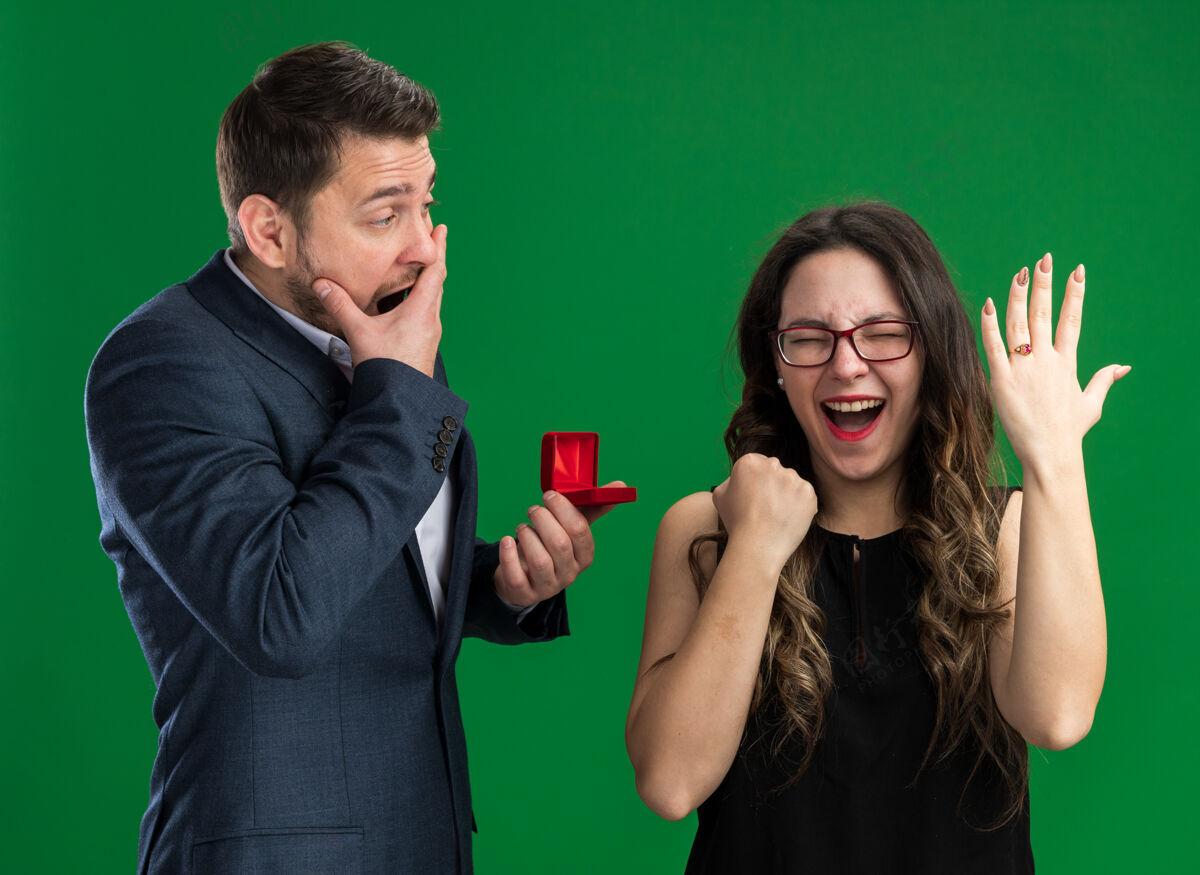 困惑年轻漂亮的夫妇困惑的男人拿着红盒子向他可爱兴奋的女友求婚 手指上戴着订婚戒指站在绿色的墙上庆祝情人节年轻举行求婚