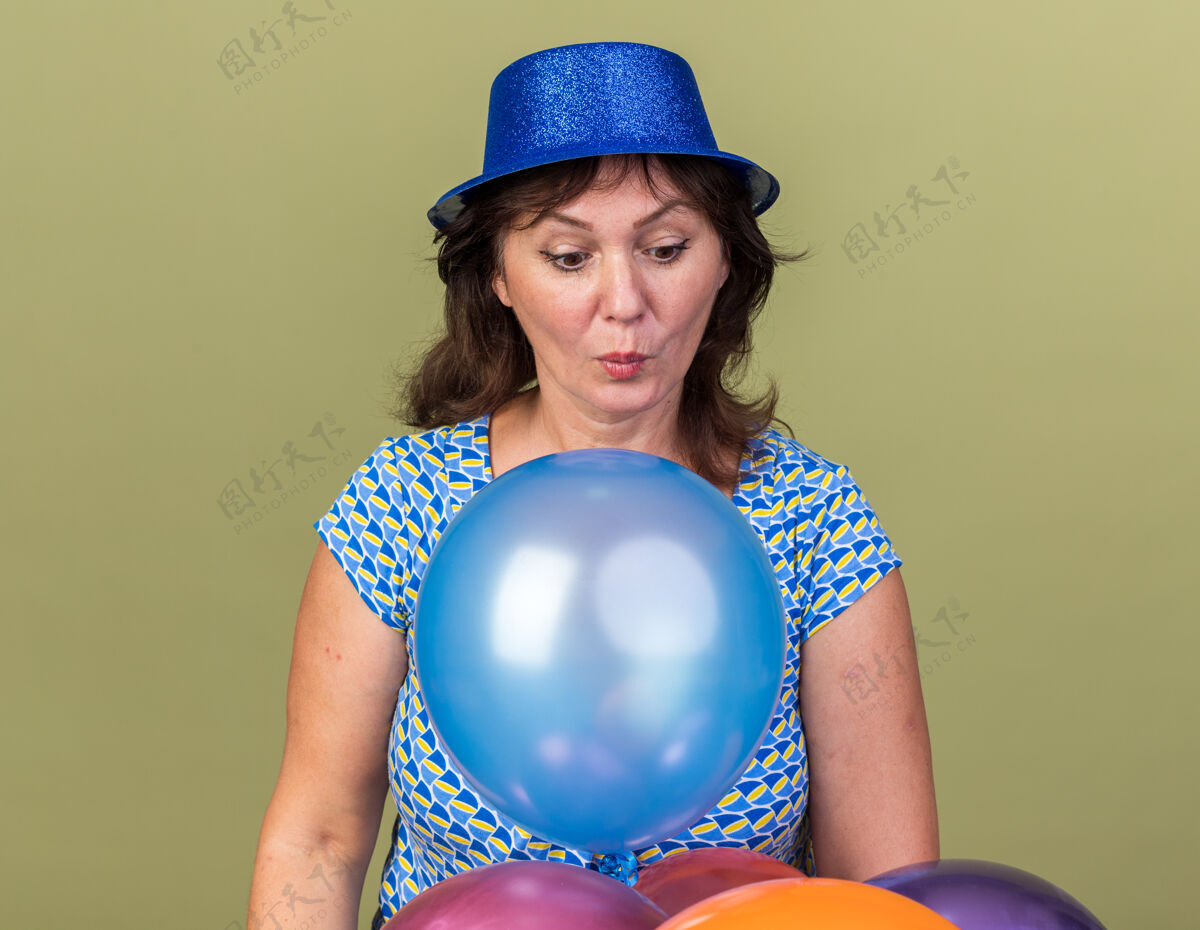 女人中年妇女戴着派对帽 手里拿着一堆五颜六色的气球 站在绿色的墙壁上庆祝生日 看起来既惊讶又惊讶帽子庆祝五颜六色