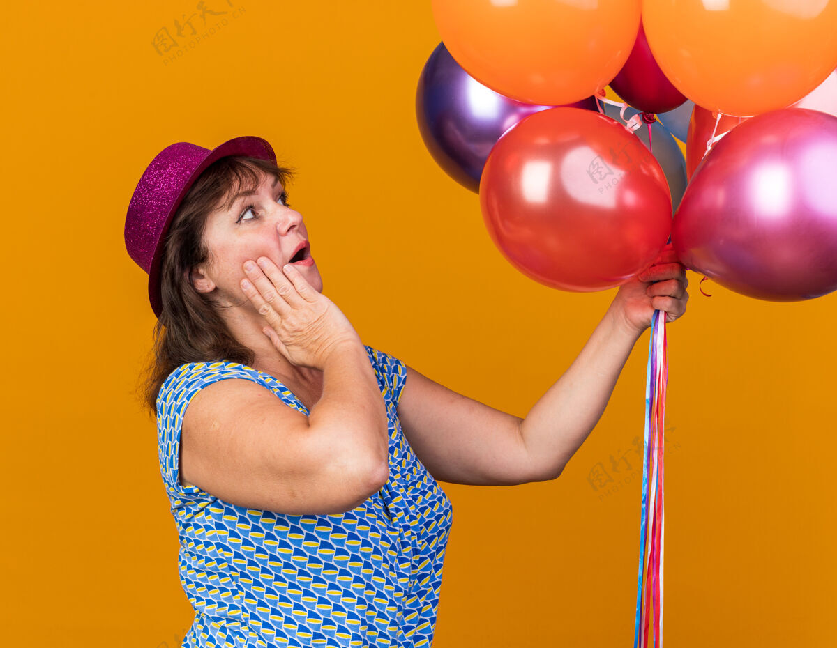 中间中年妇女戴着派对帽 手里拿着五颜六色的气球 看着他们惊讶地站在橙色的墙上庆祝生日派对年龄惊人帽子