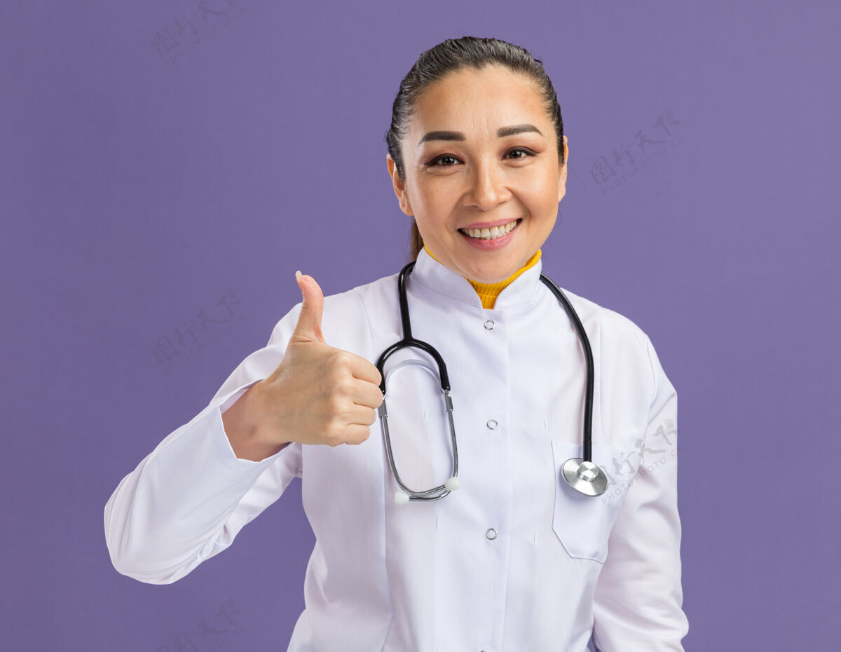 微笑身穿白色药衣 脖子上戴着听诊器的年轻女医生微笑着自信地竖起大拇指站在紫色的墙上医生脖子信心