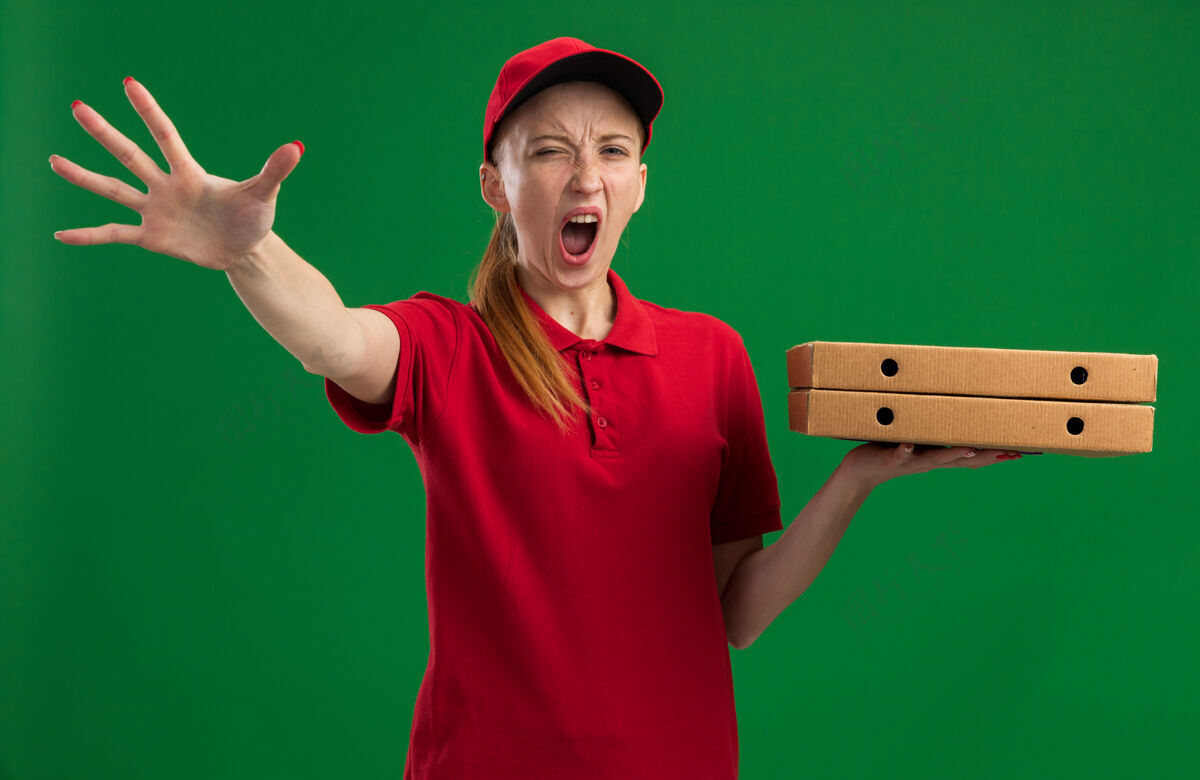 帽子身穿红色制服 头戴鸭舌帽的年轻送货员拿着披萨盒 用咄咄逼人的表情喊叫着 用手站在绿色的墙上做着停车的手势喊着手势制服