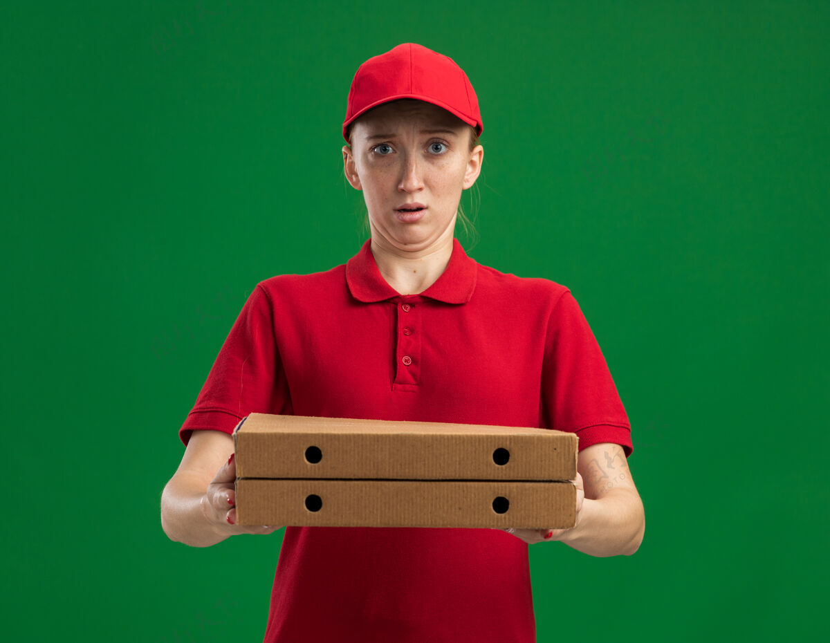 盒子身穿红色制服 头戴鸭舌帽 拿着披萨盒的年轻送货女孩站在绿色的墙上 困惑而惊讶惊喜年轻拿着