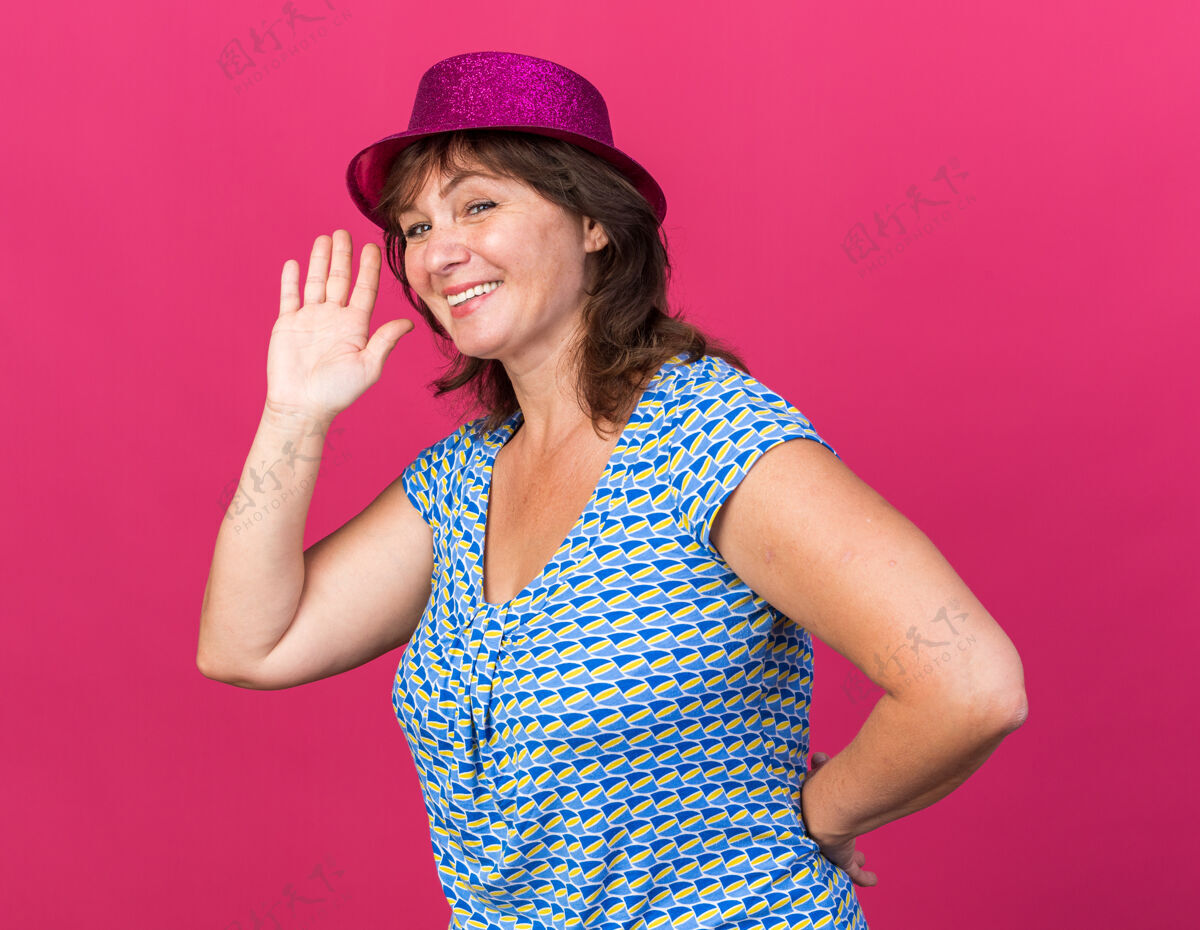 年龄快乐的中年妇女戴着派对帽 微笑着欢快地挥手庆祝生日派对站在粉红色的墙上微笑庆祝中间
