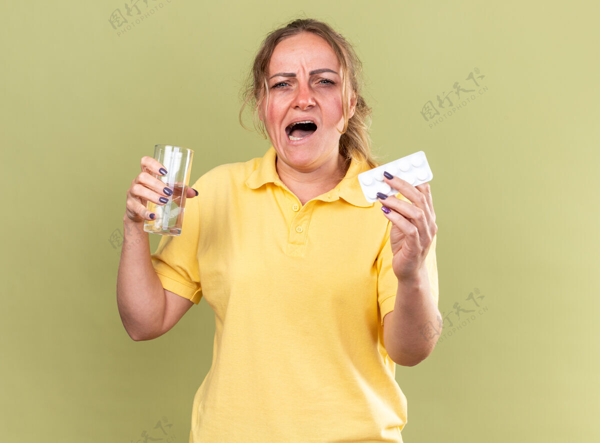 抱着一个穿着黄色衬衫的不健康的女人站在绿色的墙上 手里拿着一杯水和感冒药 感觉很糟糕站着感冒不健康