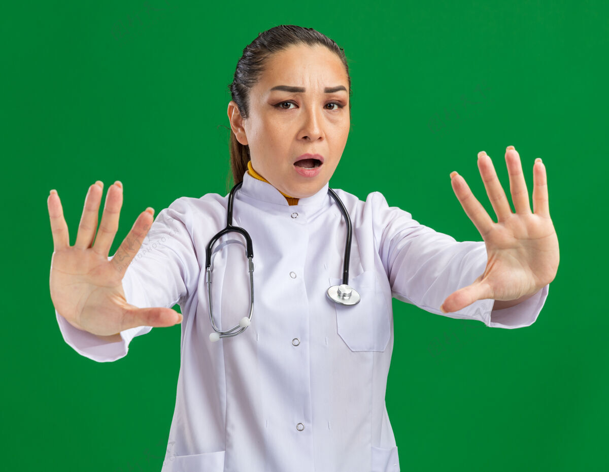 女人年轻的女医生穿着白药衣 脖子上戴着听诊器 双手站在绿色的墙上 担心地做着停止的手势停止听诊器使