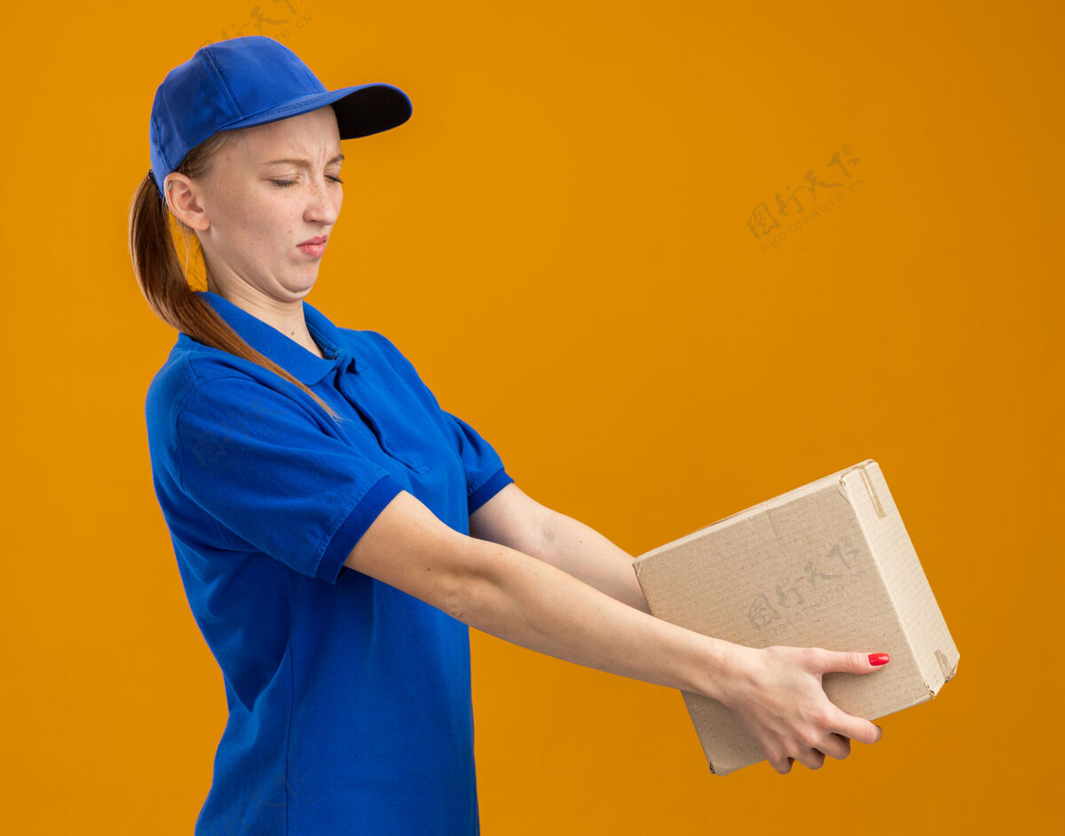 制服身穿蓝色制服 戴着帽子 拿着纸板箱的年轻送货女孩站在橙色的墙上 带着厌恶的表情看着它恶心站着表情