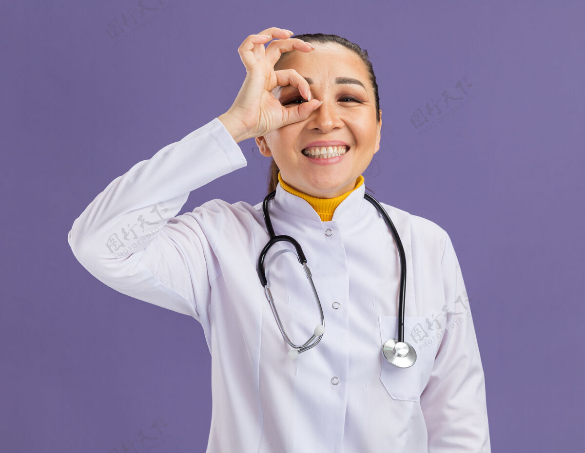 药物年轻的女医生 穿着白药衣 脖子上戴着听诊器 做着“ok”的手势 站在紫色的墙上微笑着看着这个手势脖子穿过外套