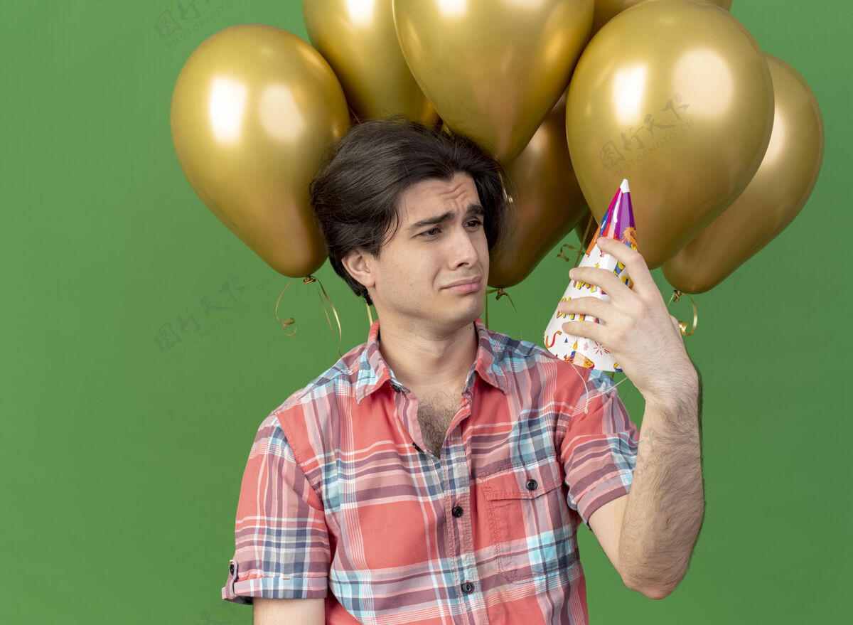 复制帅气的白种人站在氦气球前拿着生日帽看着帅气绿色氦