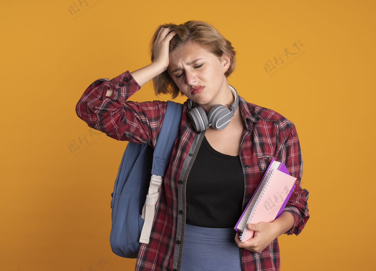 复制带着耳机 背着背包 手放在额头上 手里拿着书和笔记本的年轻斯拉夫女学生笔记本橙色学生